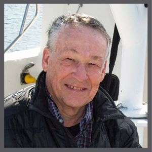 An older man with short gray hair and a black jacket smiles while sitting on a boat, with water and parts of the boat visible in the background.