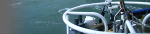 A close-up view of scientific oceanographic equipment, partially framed by white metal bars, being deployed over the side of a vessel above green, choppy seawater during Clifton Buck’s research expedition.