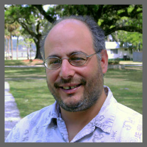 A smiling man with glasses, a receding hairline, and a short beard stands outdoors in a park with trees and a white building in the background. He is wearing a light-colored, patterned shirt.