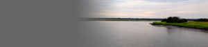 A wide river curves gently through grassy marshland under a cloudy sky, with green trees and fields visible in the distance; the left side of the image fades into a gray gradient.