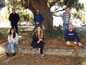 Two women and one man are sitting on a retaining wall in front of three men. They are all beneath a large live oak tree with a river view in the background.