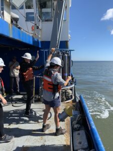 Students in hard hats and life jackets stand near the starboard rail of a research ship. The water surface is visible to the right. The students are holding on to a carousel rosette that is being maneuvered over the side of the ship.