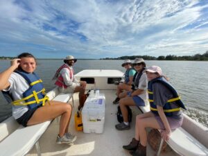 Five students wearing life jackets are sitting aboard a skiff. One male and one female student site on the port side and three female students sit on the starboard side. There is a white cooler between the groups.