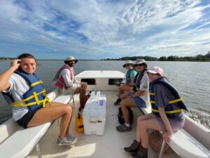 Five students wearing life jackets are sitting aboard a skiff. One male and one female student site on the port side and three female students sit on the starboard side. There is a white cooler between the groups. 