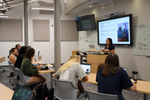 A woman stands at the front of a classroom during Semester at Skidaway, giving a presentation on Sampling Concepts to students seated at desks with laptops. A large screen displays her slides, and some students take notes.