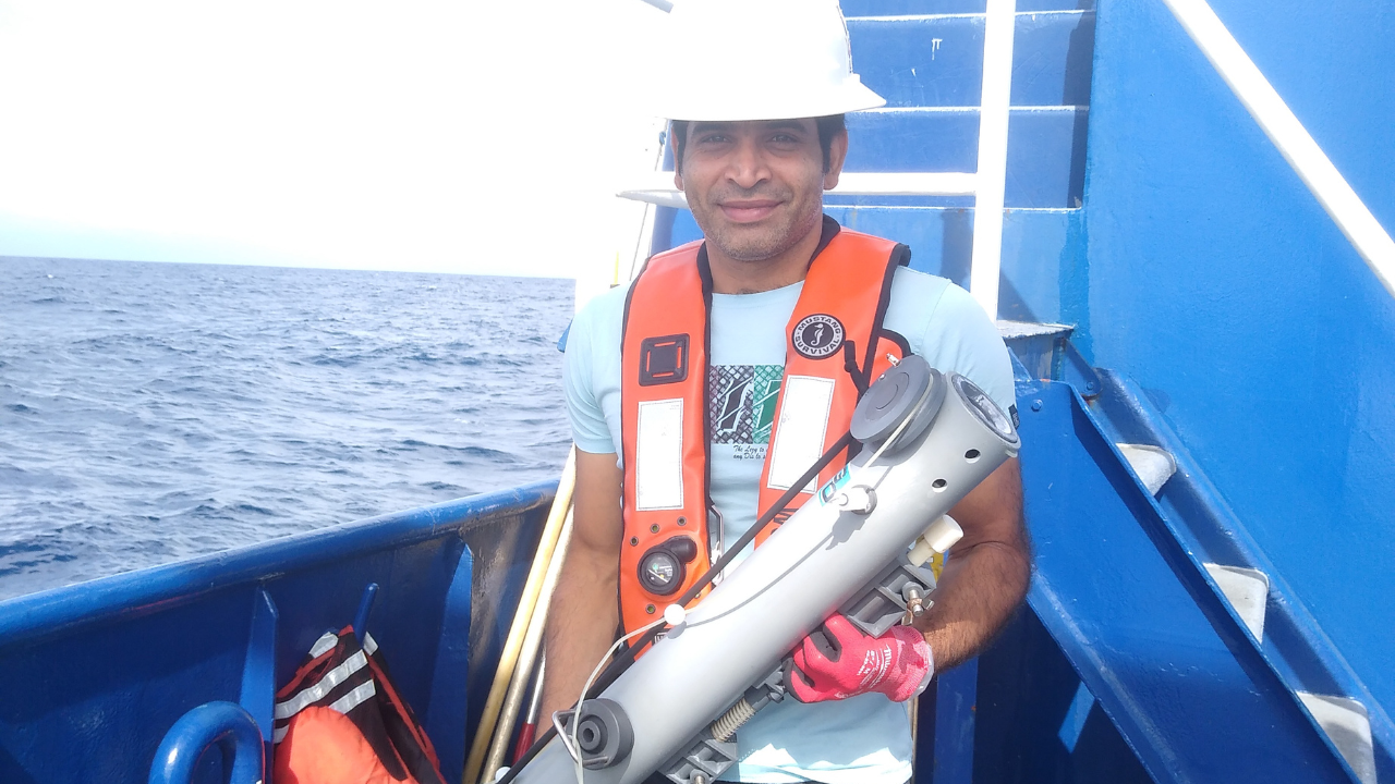 Dev Mallick, wearing a life jacket and hard hat, holds a water sampling bottle aboard the RV Savannah.