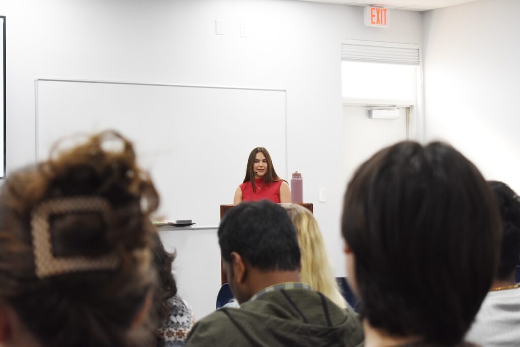 Ashley Ohall stands in an auditorium behind a podium. She's wearing a red shirt. 