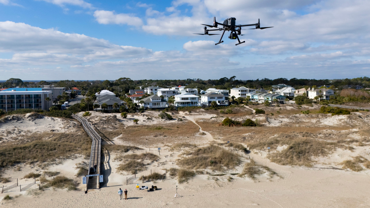 A black drone in the air over the beach. Two researchers stand observing the drone.