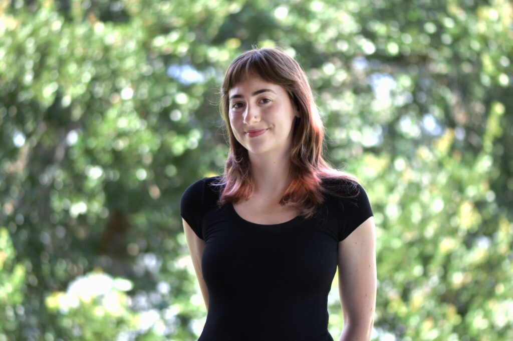 A young woman with light skin and shoulder-length brown hair stands outdoors, smiling slightly. She is wearing a short-sleeved black top, with a blurred background of green trees and sunlight.