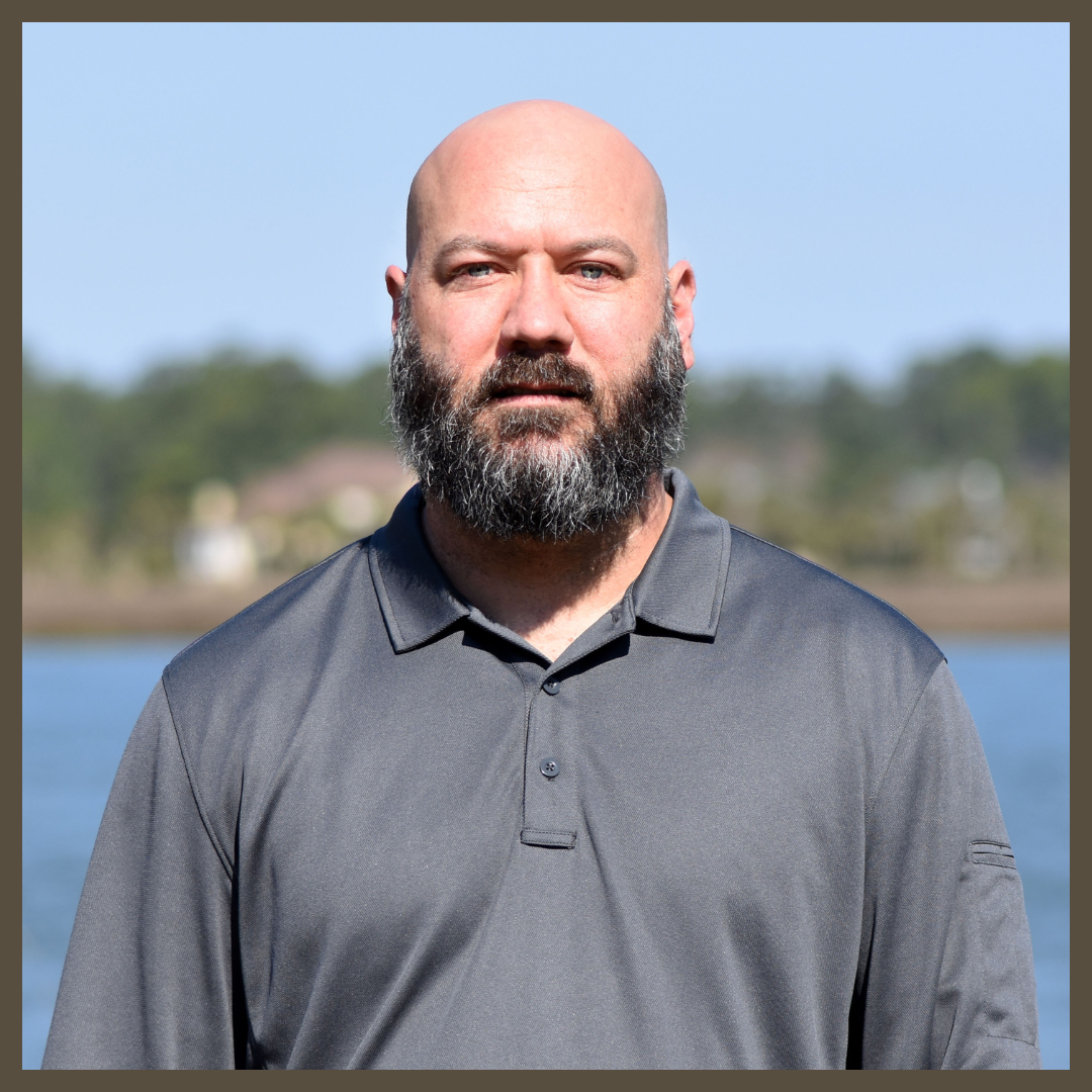A bald man with a thick beard, wearing a gray polo shirt, stands outdoors in front of a body of water with trees and buildings blurred in the background.