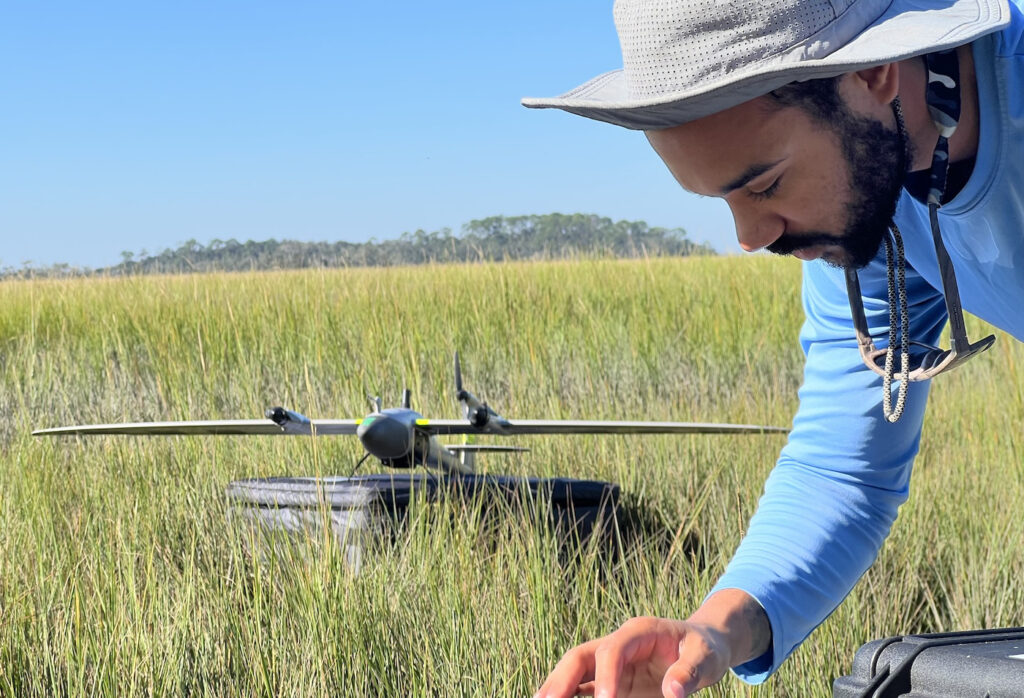 A person in a light blue shirt and wide-brimmed hat works in a grassy field with a drone resting on a black case in the background under a clear blue sky.