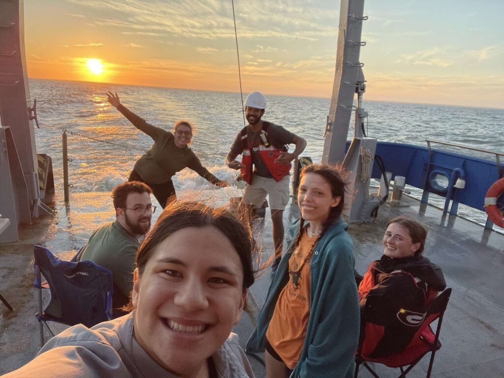Six people smile and pose on a boat at sunset, with the sun low over the ocean. Some sit on folding chairs while others stand, including one in a hard hat and life jacket. The scene is relaxed and cheerful.