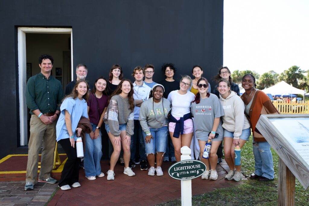A group of sixteen people poses and smiles outside a black building near a sign reading Lighthouse. The group includes both men and women, some standing and some crouching, with trees and tents visible in the background.