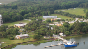 Aerial view of a waterfront research facility with several buildings surrounded by trees, a dock with a blue and white boat, and a large grassy area in the background.