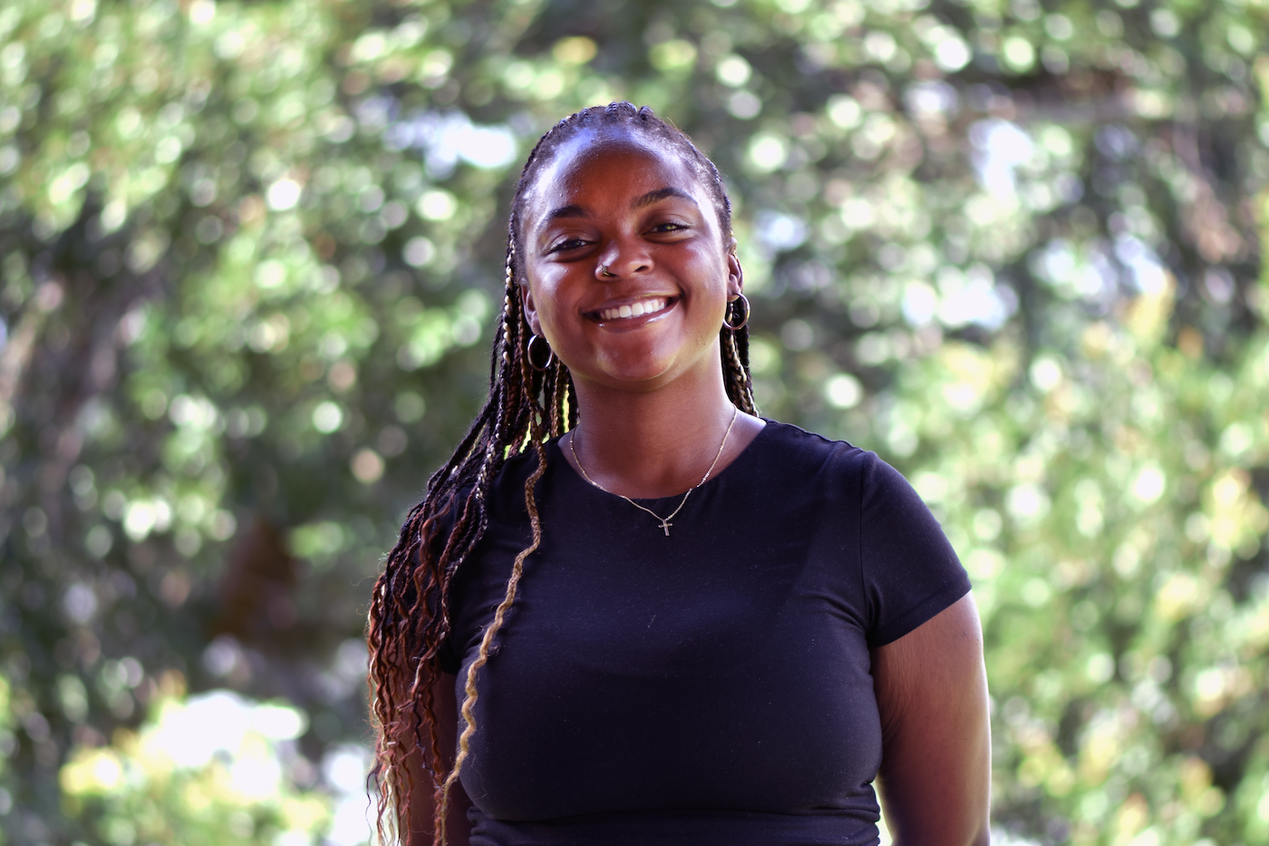 A woman with long braids, wearing a black t-shirt and a necklace with a cross, stands outdoors smiling in front of a background of green, sunlit trees.