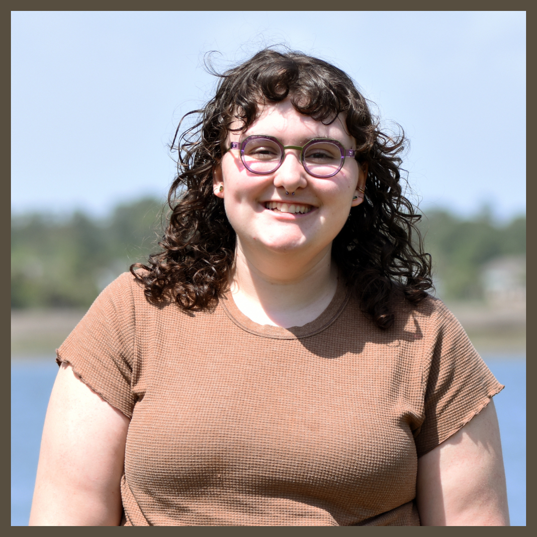A person with curly brown hair and glasses smiles outdoors. They are wearing a brown short-sleeved top. The background is blurred, showing water and green trees under a blue sky.