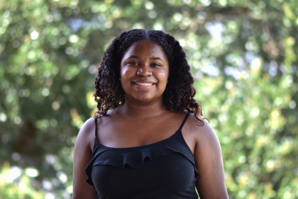 A woman with curly hair, wearing a black sleeveless top, smiles while standing outdoors with green trees blurred in the background.