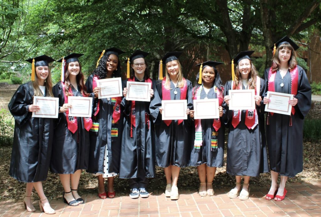 Eight graduates in caps and gowns stand outside on a brick path, holding certificates and smiling. Green trees and sunlight are in the background, creating a cheerful, celebratory atmosphere.