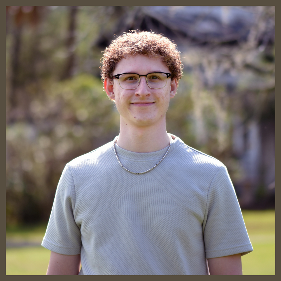 A young person with curly hair and glasses, wearing a light gray textured shirt and a chain necklace, standing outside with greenery and blurred trees in the background.