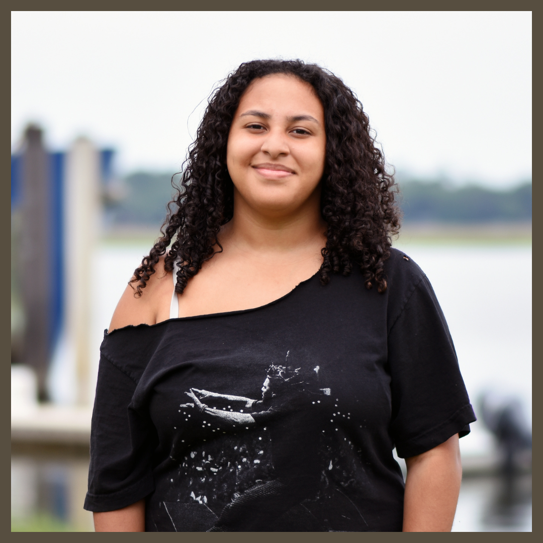 A young woman with curly hair smiles at the camera, wearing a black off-the-shoulder T-shirt. She stands outdoors near a body of water with blurred docks and greenery in the background.