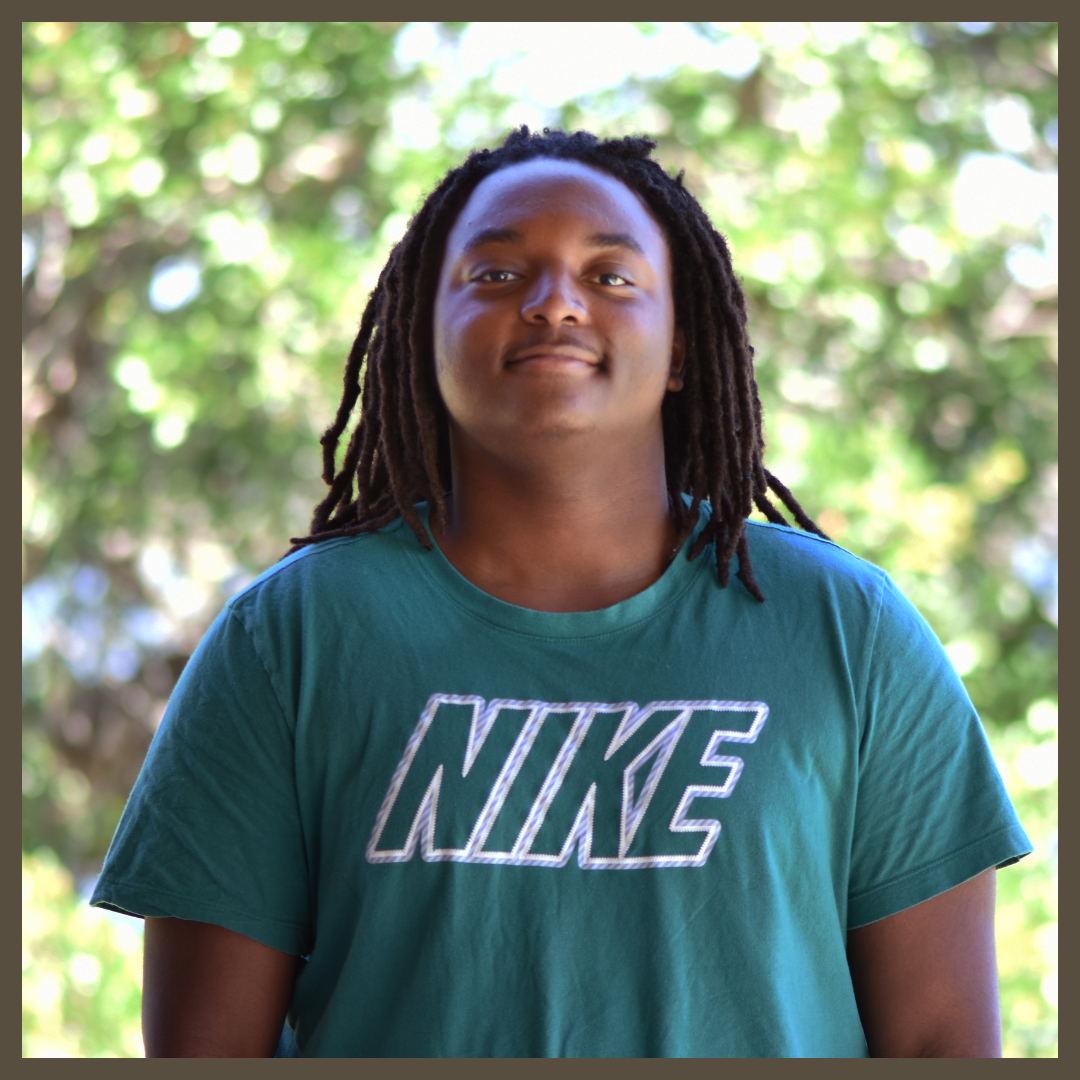 A person with long dreadlocks wears a green Nike t-shirt and stands outdoors, smiling slightly. The background is blurred with green foliage and sunlight.