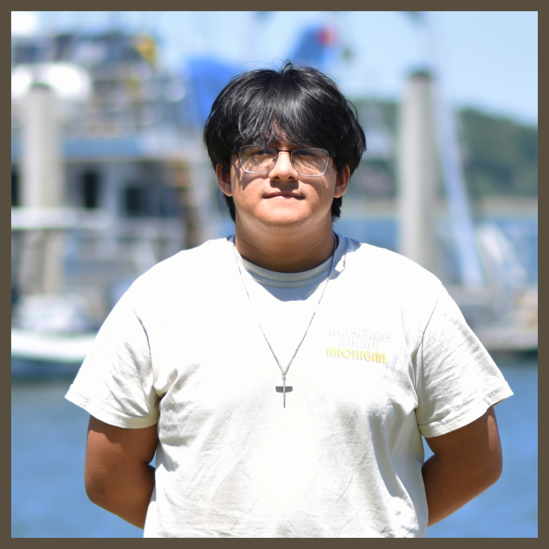 A person with dark hair and glasses wearing a light t-shirt and a necklace with a cross stands outdoors in front of blurred boats on the water.