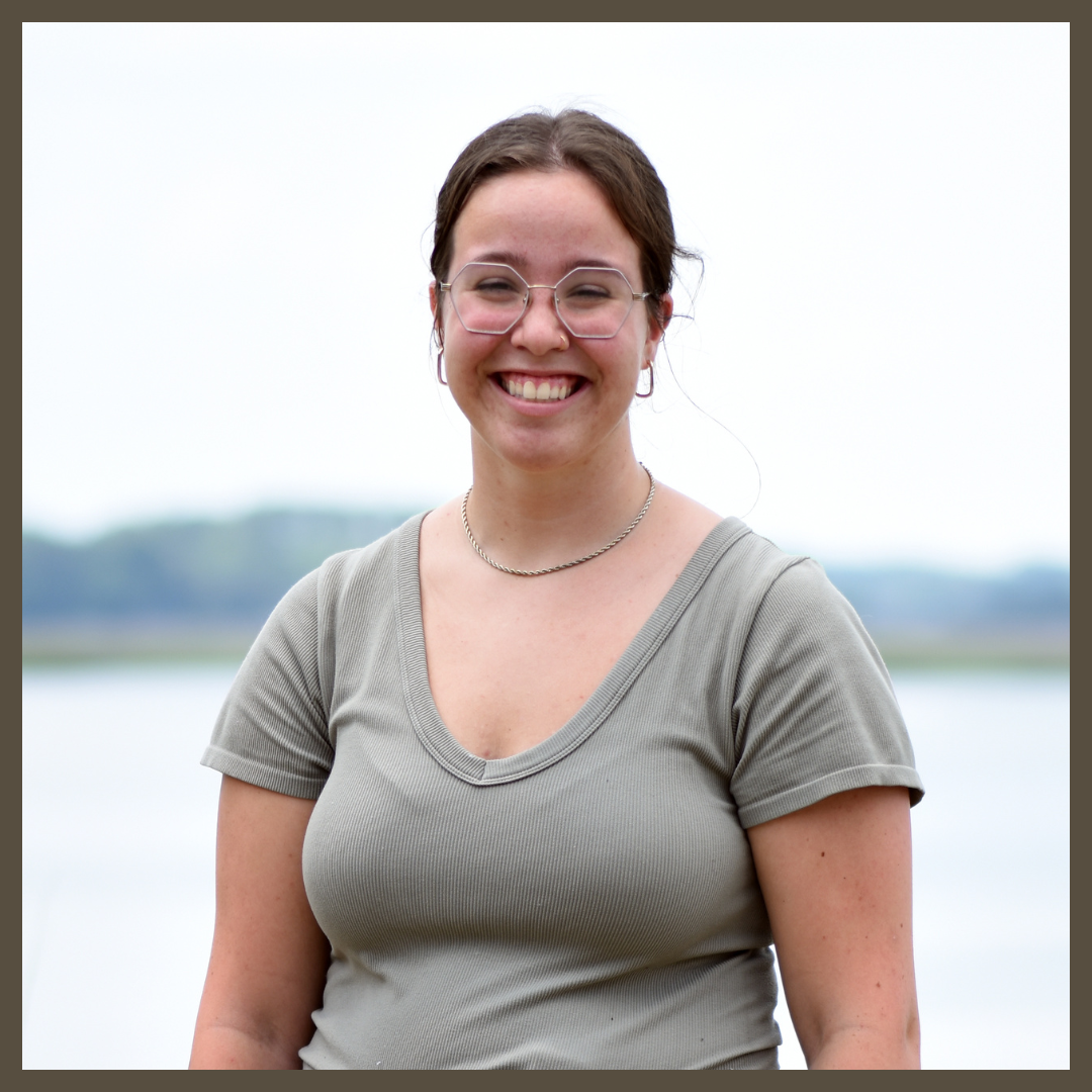 A young woman with glasses and brown hair tied back smiles at the camera. She is wearing a light gray short-sleeved shirt and a necklace, standing outdoors with water and blurred greenery in the background.