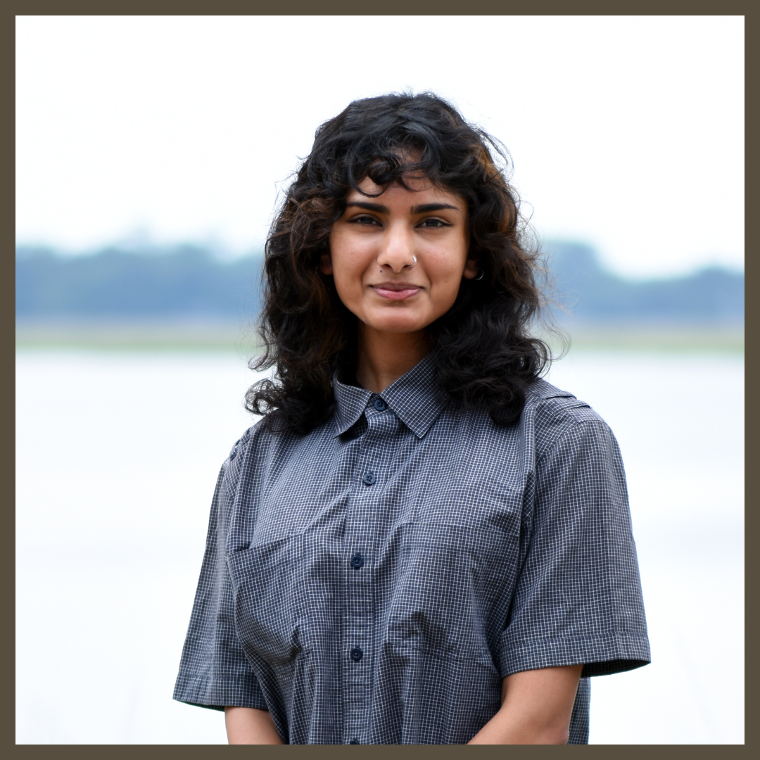 A young woman with wavy dark hair, wearing a short-sleeved gray checkered shirt, stands outdoors smiling gently with a blurred water and tree background.