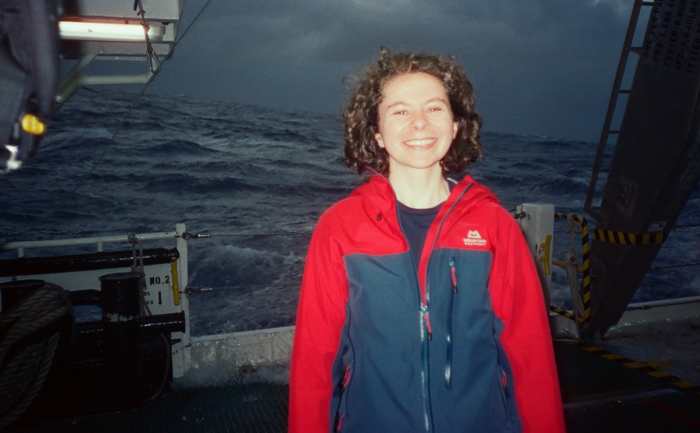 A person with curly hair and a red jacket smiles on a ship’s deck with the ocean and cloudy sky in the background.