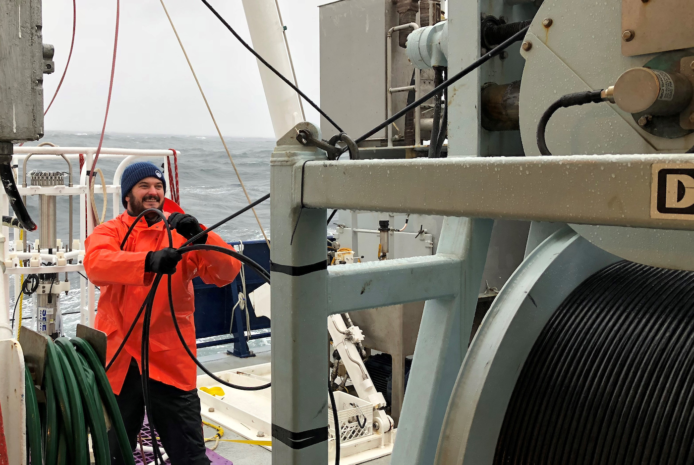 A person in an orange raincoat and blue hat works with thick cables on a ship deck, surrounded by equipment, with the ocean and cloudy sky in the background.