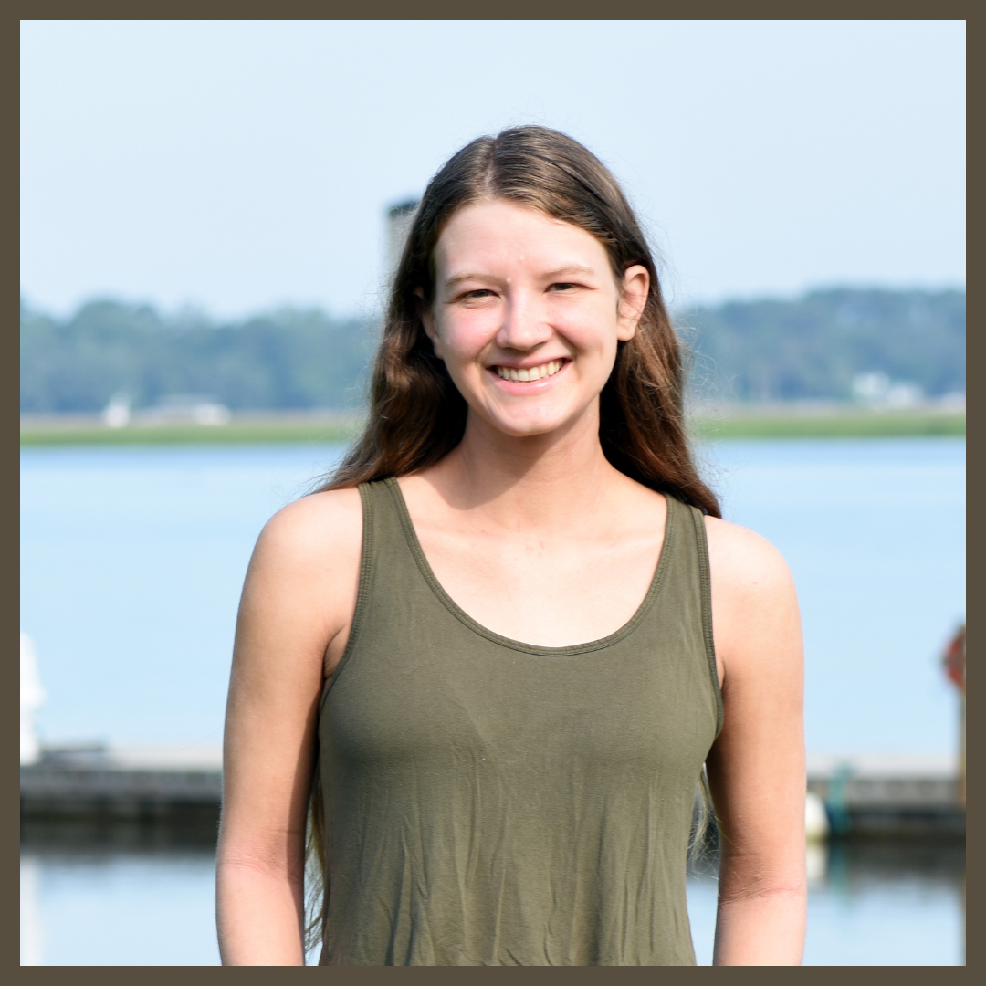 A young woman with long brown hair, wearing an olive green sleeveless top, smiles while standing outdoors by a body of water with a dock and trees in the background.