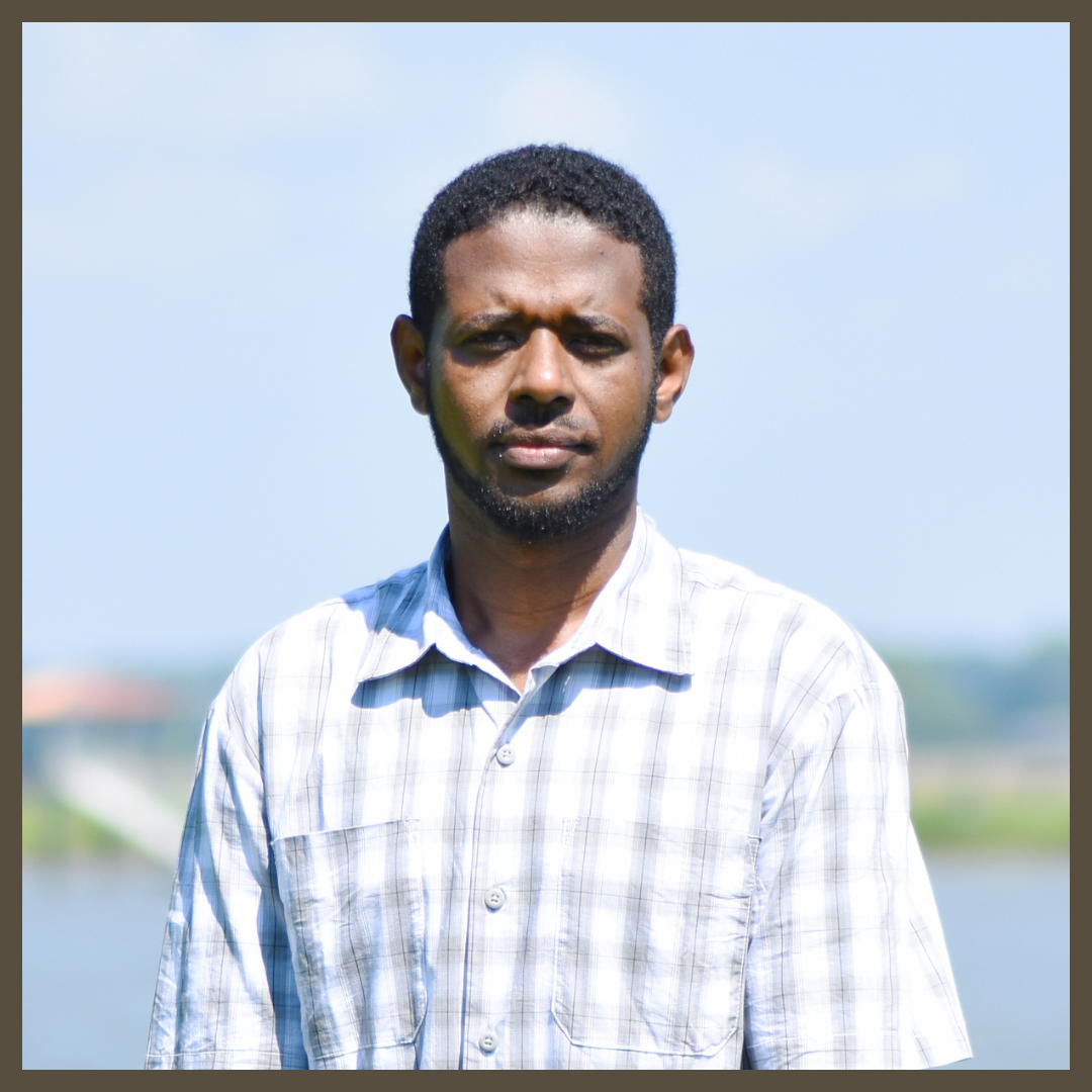 A man wearing a light plaid shirt stands outdoors in sunlight with a blurred background of water and sky.