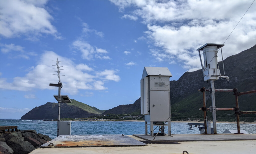 Weather and communication equipment are installed on a concrete pier by the ocean, with green mountains and a partly cloudy sky in the background.