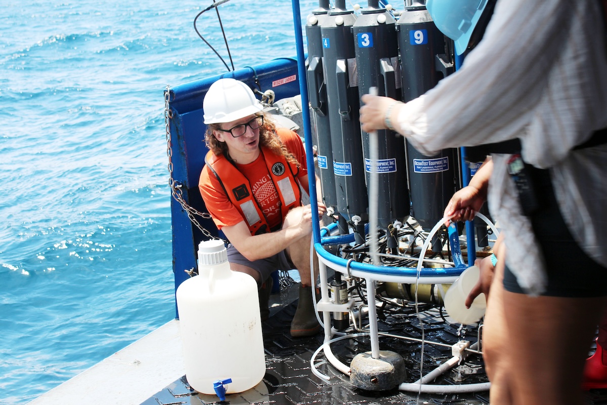 A person in an orange shirt and hard hat kneels on a boat beside scientific equipment, while another person in a blue helmet stands nearby. The ocean is visible in the background.