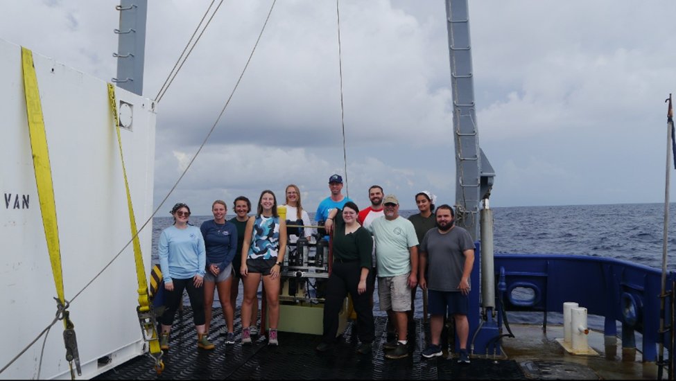 Ten people stand together on the deck of a research vessel at sea, posing in front of equipment under a cloudy sky. The ocean is visible in the background, and the group appears to be part of a marine research expedition.