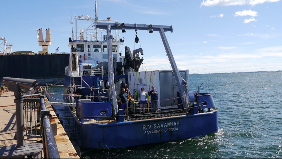 A blue and white research vessel named R/V Savannah is docked at a pier. Several people are standing on its deck, and the sea and sky are visible in the background.