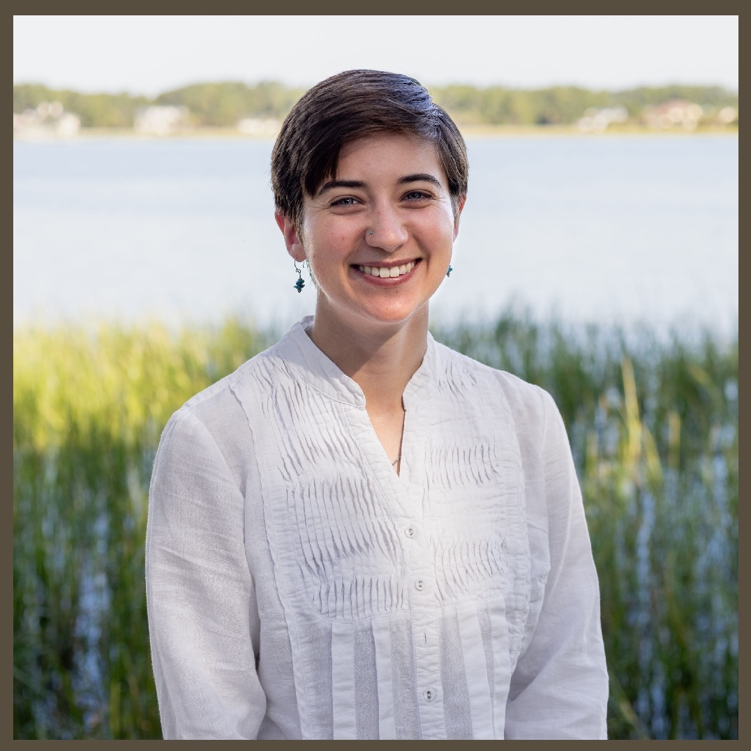 A person with short brown hair and a white shirt stands smiling in front of a lake, with green grass and trees in the background on a sunny day.